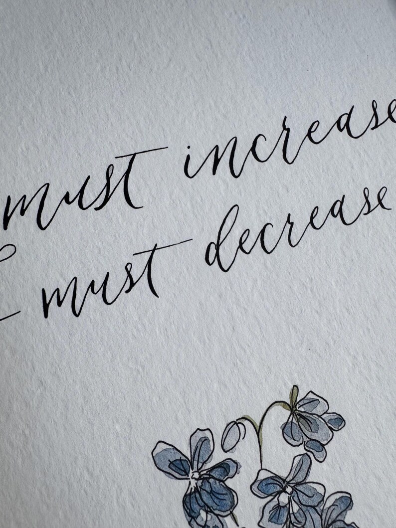 May include: A white textured paper featuring black calligraphy text that reads "must increase" and "I must decrease" positioned above a hand-drawn illustration of small blue flowers with green stems and leaves rendered in a watercolor style.