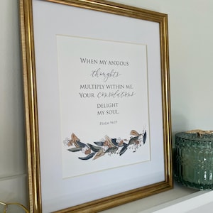 May include: A framed print with a gold frame and a white mat. The print features a black and white calligraphy quote with the text "WHEN MY ANXIOUS thoughts MULTIPLY WITHIN ME, YOUR Consolations DELIGHT MY SOUL. PSALM 94:19". The quote is surrounded by a wreath of watercolor leaves and flowers.
