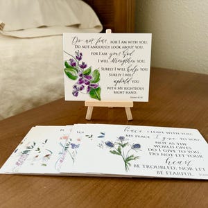May include: Set of six inspirational cards with floral illustrations and bible verses. The cards feature a watercolor style with purple and blue flowers. The text on the cards includes verses from the Bible, such as "Do not fear, for I am with you" and "Peace I leave with you; my peace I give to you."