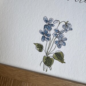 May include: A white paper print featuring a watercolor illustration of blue violet flowers with green leaves and stems. The text "I must decre" is visible in black cursive script at the top. The print is housed in a light brown wooden frame.