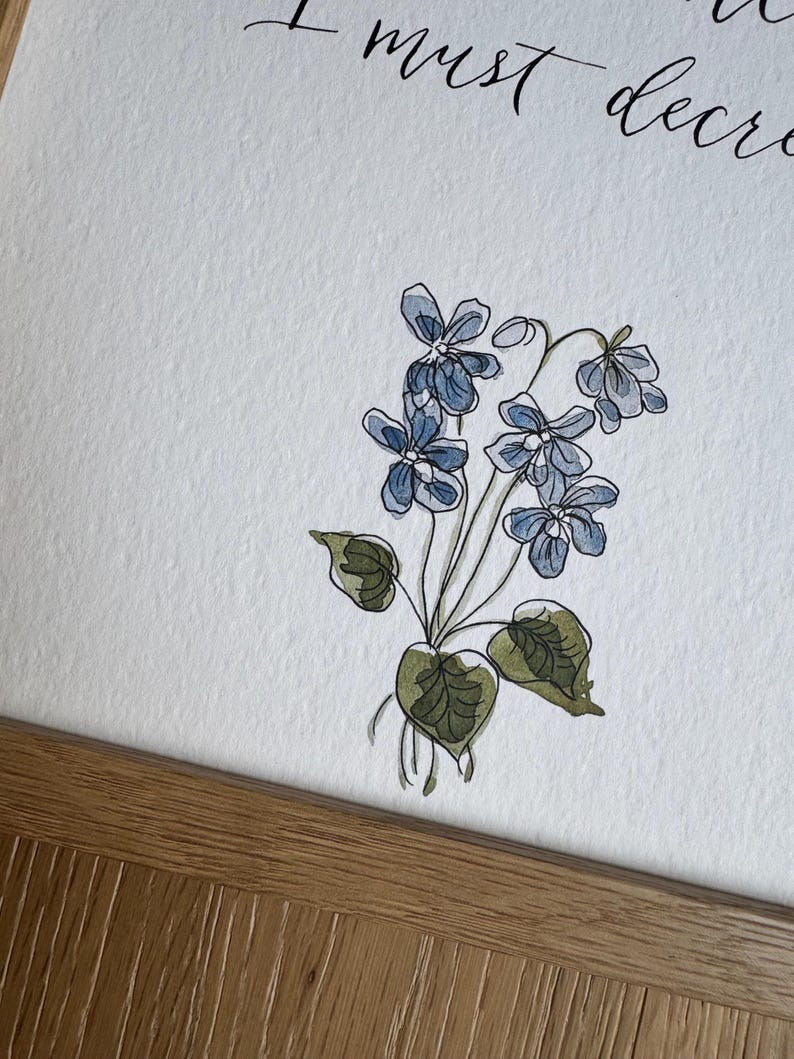 May include: A white paper print featuring a watercolor illustration of blue violet flowers with green leaves and stems. The text "I must decre" is visible in black cursive script at the top. The print is housed in a light brown wooden frame.