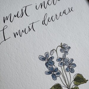 May include: A white textured paper print featuring black calligraphy that reads, "must increase. I must decrease." Below the text is a watercolor illustration of a cluster of small blue violet flowers with green leaves and thin stems.