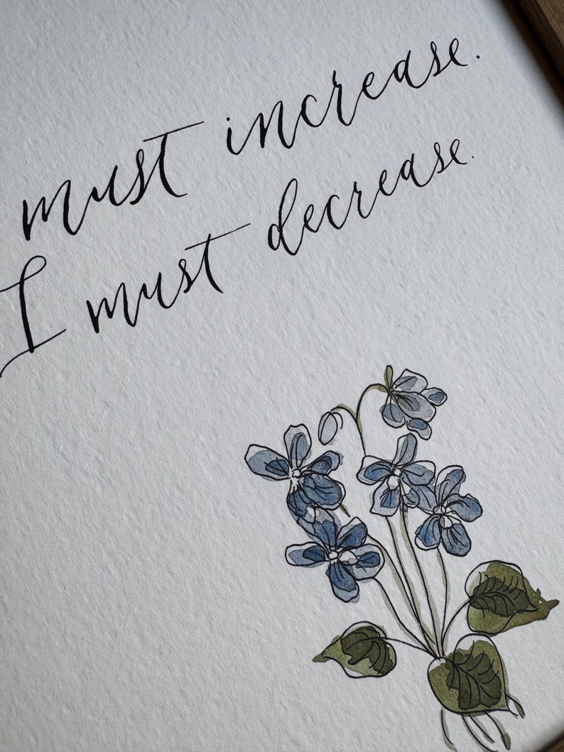 May include: A white textured paper print featuring black calligraphy that reads, "must increase. I must decrease." Below the text is a watercolor illustration of a cluster of small blue violet flowers with green leaves and thin stems.