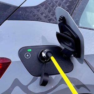 Puede incluir: Un coche eléctrico gris con un cable de carga amarillo conectado al puerto de carga. El puerto de carga está ubicado en la parte trasera del lado del pasajero del coche.