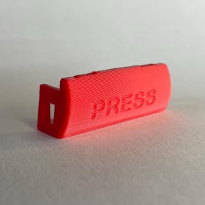 Op de afbeelding: Een felrood, 3D-geprint kunststof onderdeel met een rechthoekig, licht gebogen voorvlak waarop het woord PRESS in hoofdletters is gedrukt. Het object heeft een holle, doosachtige structuur aan één kant met een kleine vierkante opening.