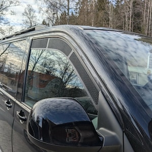 Peut inclure: Un van noir avec des vitres teintées et un porte-bagages de toit. La vitre latérale comporte un évent en maille. Le rétroviseur latéral reflète l'environnement. Le véhicule est garé à l'extérieur par temps nuageux.