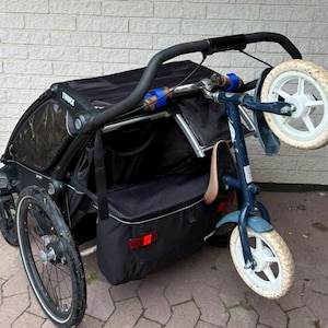 May include: A black Thule Chariot child carrier with a blue and black bike attached to the back. The carrier has a large black storage compartment and a black canopy. The bike has a white seat and white wheels.