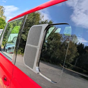 Könnte beinhalten: Seitenfenster eines roten Fahrzeugs mit einem silberfarbenen Lüftungsdeckel mit Netzmuster. Das Fenster spiegelt den Himmel und die umliegenden Bäume wider. Der Lüftungsdeckel hat eine rechteckige Form mit abgerundeten Ecken.