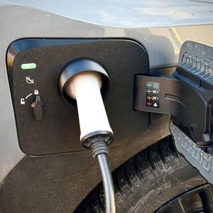 May include: Close-up of an electric vehicle charging port with a white charging cable plugged in. The port is black and has a hinged door. The car is grey, and the tyre is visible.