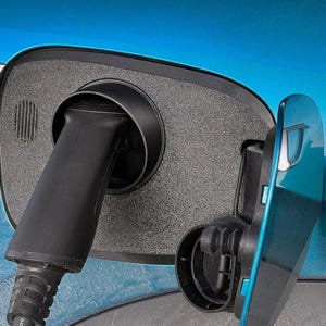 May include: Close-up of an electric vehicle charging port with a black charging cable inserted. The port is open, revealing the charging receptacle. The car's body is a bright teal color, with a silver "DESIGN" detail.