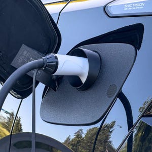 Puede incluir: Primer plano de un puerto de carga de vehículo eléctrico con un cable de carga conectado. El puerto está abierto, revelando el enchufe de carga. La carrocería del coche es negra y el puerto de carga es gris. SAIC VISION LED lighting system.