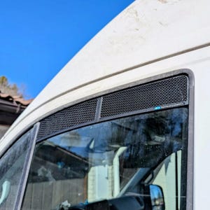 May include: A black mesh vent cover installed above a vehicle window. The vent cover is rectangular and has a fine, diamond-shaped pattern. The vehicle is white, and the sky is blue.