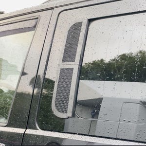 Könnte beinhalten: Nahaufnahme eines schwarzen Fahrzeugseitenfensters mit einem rechteckigen, gemusterten Netzgitter. Der Bildschirm ist silber und schwarz, und das Fenster ist mit Regentropfen bedeckt. Der Hintergrund zeigt eine verschwommene Ansicht von Bäumen und einem anderen Fahrzeug.