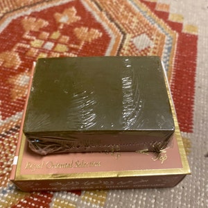 May include: A rectangular, olive-green bar of soap wrapped in clear plastic sits atop a pink and gold box. The box is labeled "Royal Oriental Selection" in gold lettering. The background is a patterned rug.
