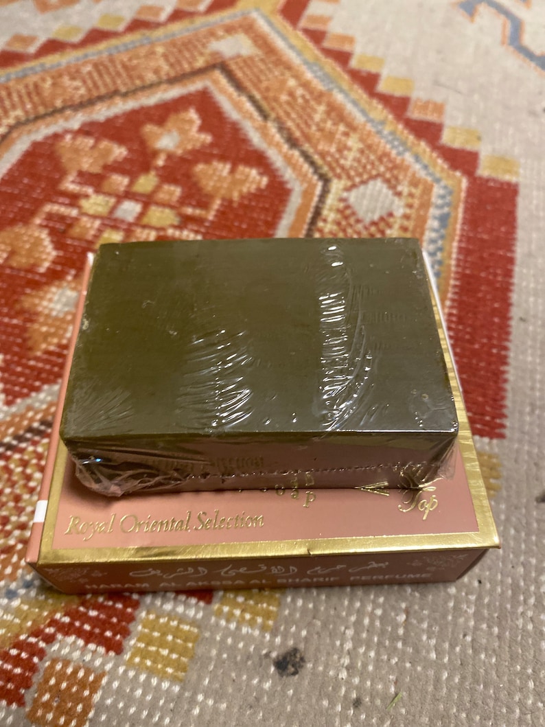 May include: A rectangular, olive-green bar of soap wrapped in clear plastic sits atop a pink and gold box. The box is labeled "Royal Oriental Selection" in gold lettering. The background is a patterned rug.