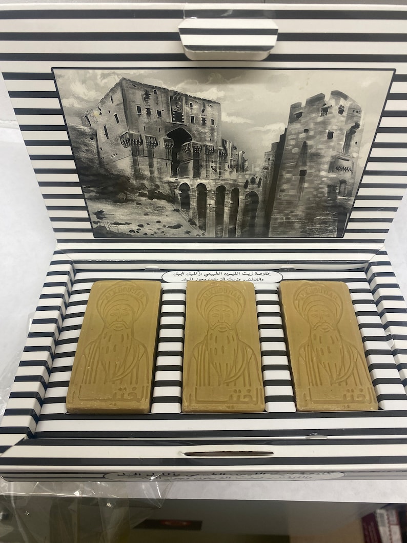 May include: Three bars of brown soap in a white and black striped box. The soap is embossed with an image of a person with a beard and turban. The box has a black and white image of a building on the lid. The text on the box reads "غالية زيت الزيتون الطبيعي" and "صابون زيت الزيتون الطبيعي".