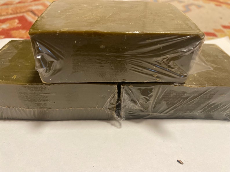 May include: Three rectangular bars of olive-green soap are stacked on top of each other, wrapped in clear plastic. The soap has a slightly uneven surface, and the packaging is tightly sealed. The bars are arranged on a white surface.