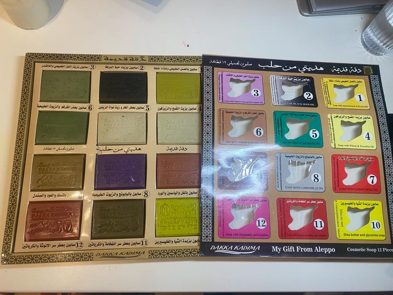 Op de afbeelding: Een set van 12 repen Aleppo-zeep in een doos met een gouden rand. De zeep is verpakt in afzonderlijke vierkanten met Arabische tekst en nummers. De doos heeft de tekst "Dakka Kadima My Gift From Aleppo Cosmetic Soap 12 Piece".