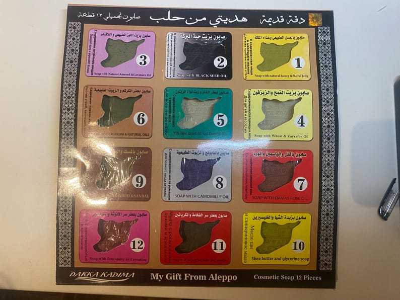 Op de afbeelding: Een set van 12 kleine zeepjes, elk in de vorm van de omtrek van Syri&euml;. De zeepjes zijn gerangschikt in een raster van vier rijen en drie kolommen. Elk zeepje heeft een nummer van 1 tot 12 en een beschrijving van de ingredi&euml;nten van de zeep in het Arabisch. De set is gelabeld met "My Gift From Aleppo" en "Cosmetische Zeep 12 Stuks".