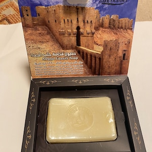 May include: A bar of Aleppo Luxury Soap in a decorative box. The box features an illustration of a stone fortress and text in multiple languages. The soap is rectangular and has a circular emblem. The box is dark with gold accents.