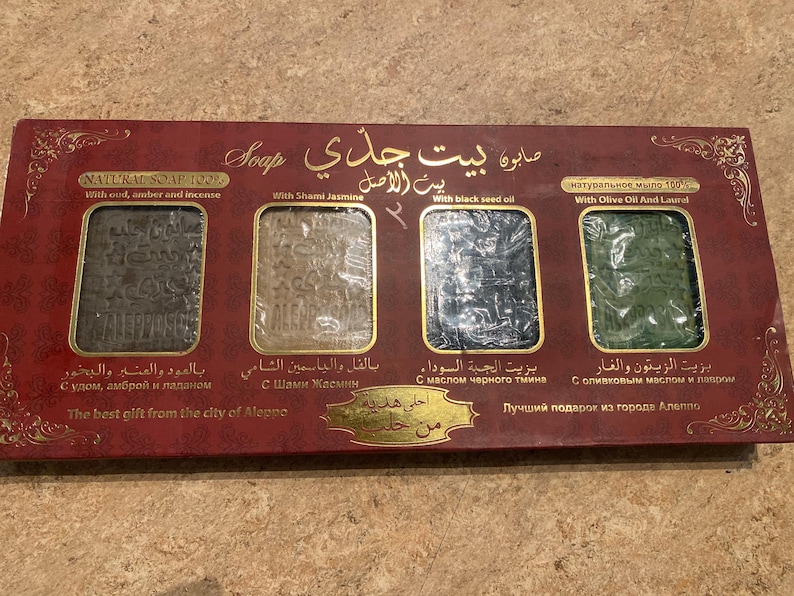 May include: A red box with four bars of soap. The soap bars are labeled with Arabic text and English translations. The text reads "Natural Soap 120%" with oud, amber, and incense, "With Shami Jasmine", "With black seed oil", and "Natural soap 100% with Olive Oil and Laurel". The box also says "The best gift from the city of Aleppo".