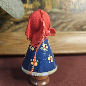 Może przedstawiać: Drewniana figurka osoby w czerwonym kapturze i niebieskiej sukience ozdobionej ż&oacute;łtymi i białymi kwiatowymi wzorami. Figurka ma brązową podstawę i jasnoniebieskie wykończenie.