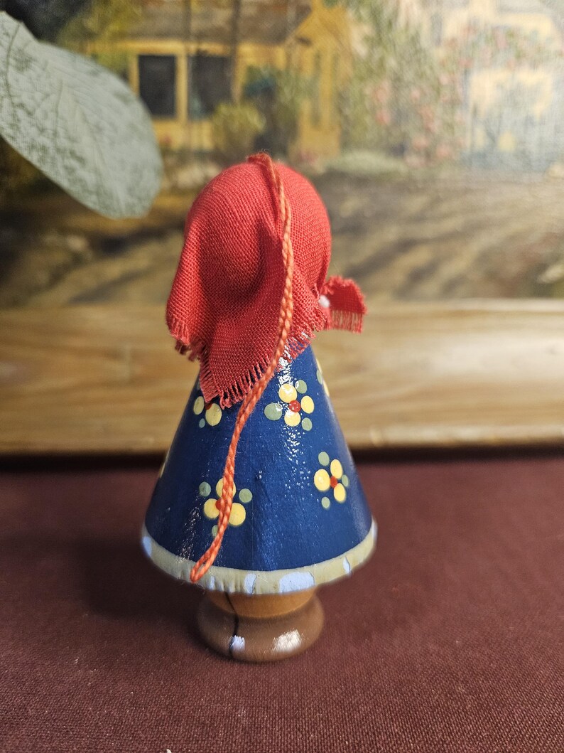 Może przedstawiać: Drewniana figurka osoby w czerwonym kapturze i niebieskiej sukience ozdobionej ż&oacute;łtymi i białymi kwiatowymi wzorami. Figurka ma brązową podstawę i jasnoniebieskie wykończenie.