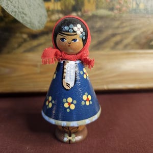 Może przedstawiać: Drewniana lalka z namalowaną twarzą, w czerwonej chuście i niebieskiej sukience ozdobionej ż&oacute;łtymi i czerwonymi kwiatami. Lalka ma czarne nakrycie głowy i stoi na brązowej podstawie. Wysokość ok. 10 cm.