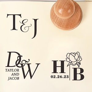May include: Wooden stamps with round handles and circular bases are arranged near a notebook with custom text. The notebook displays initials and names, including "T&J", "D&W TAYLOR AND JACOB", and "HB 02.26.23" with a floral design. A white flower is in the background.
