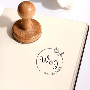 May include: A wooden stamp with a round handle and base, on a cream notebook. The stamp features a floral design with the initials "W&J" and the date "04-05-2025" within a circle. A white tulip and two black pens with gold detailing are also visible.