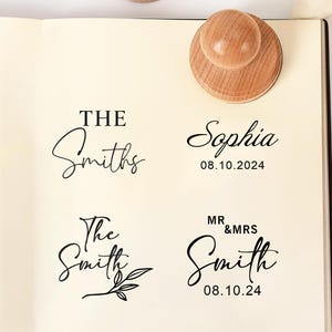 May include: Four round wooden stamps with light brown grain patterns are placed on a page showing black ink impressions. The prints display text including The Smiths, Sophia 08.10.2024, The Smith with a leaf graphic, and MR & MRS Smith 08.10.24.