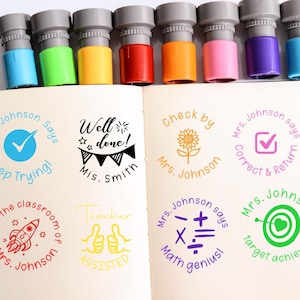 Pode incluir: Um conjunto de carimbos coloridos para professores com tampas cinzentas e bases de várias cores. Os carimbos incluem frases como "Keep Trying", "Well Done" e "Target Achieved". Os carimbos estão numa página de caderno branca.