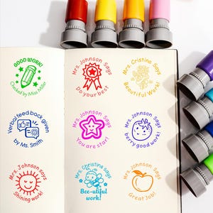 May include: A collection of colorful self-inking stamps with various designs and phrases, including "Good Work!" and "Do your best." The stamps have red, yellow, orange, and pink tops. The stamps are next to a notebook with stamped images.