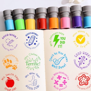 Op de afbeelding: Een set kleurrijke zelfinktende stempels met grijze bovenkanten en verschillende gekleurde bases. De stempels zijn gerangschikt boven een open notitieboek met positieve berichten zoals "Good Work!" en "You Did It!" in verschillende lettertypen en kleuren.