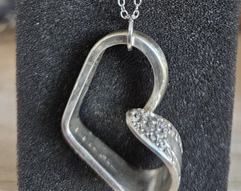 May include: A silver heart pendant necklace. The heart-shaped pendant is crafted from repurposed silverware and features a floral design on one side. The necklace hangs from a delicate silver chain, showcasing a unique, handcrafted jewelry piece.