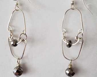 May include: A pair of silver-toned dangle earrings. Each earring features a geometric design with a faceted, dark gray bead at the bottom and a smaller bead in the middle. The earrings hang from a silver hook.