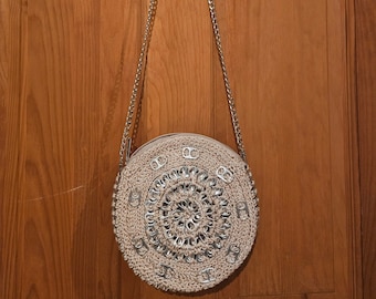 May include: A round, beige crocheted handbag with a silver chain strap. The bag's center features a circular design made from silver pull tabs. The bag is hanging against a wooden door.