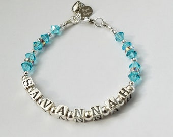 May include: A silver bracelet with the name "SAVANNAH" spelled out in silver beads. The bracelet is accented with light blue crystal beads.