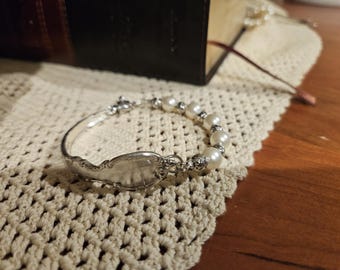 May include: A bracelet featuring a curved silver-toned metal handle segment from a spoon, connected to a strand of white spherical beads separated by small, ornate silver-toned metal spacers, forming a circular jewelry piece.