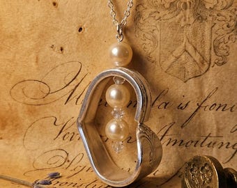 May include: A silver-toned metal heart-shaped pendant suspended from a delicate chain. The pendant features three cream-colored spherical beads and two small clear faceted beads arranged vertically within the center of the heart frame.