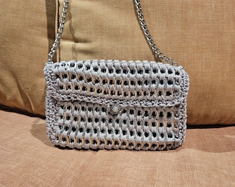 May include: A silver chain strap purse made from woven metal rings. The purse is sitting on a brown fabric surface.