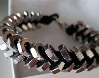 May include: A bracelet constructed from a series of silver-toned hexagonal metal nuts threaded onto a black cord. The metallic nuts are arranged in a repeating pattern along the length of the cord, which features a metal clasp closure at the end.