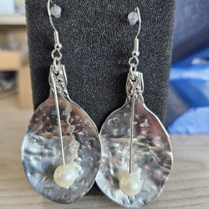 May include: A pair of silver-toned, hammered metal earrings. Each earring has a teardrop shape with a textured surface and a small, round, off-white pearl bead. The earrings hang from silver-colored ear wires.