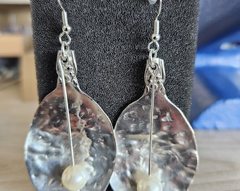May include: A pair of silver-toned, hammered metal earrings. Each earring has a teardrop shape with a textured surface and a small, round, off-white pearl bead. The earrings hang from silver-colored ear wires.