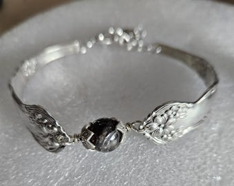 May include: Silver bracelet crafted from repurposed silverware. The bracelet features ornate floral and scroll designs on the spoon heads, connected by a dark, round gemstone. A unique, handcrafted jewelry piece.