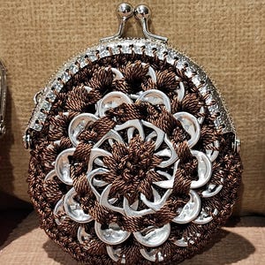 May include: A handmade coin purse with a silver clasp and a unique design. The purse is crafted from brown yarn and recycled aluminum can tabs, arranged in a floral pattern. The purse has a rounded shape.