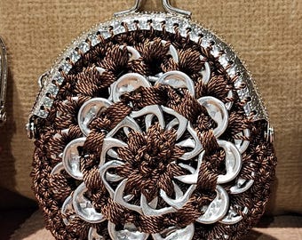 May include: A handmade coin purse with a silver clasp and a unique design. The purse is crafted from brown yarn and recycled aluminum can tabs, arranged in a floral pattern. The purse has a rounded shape.
