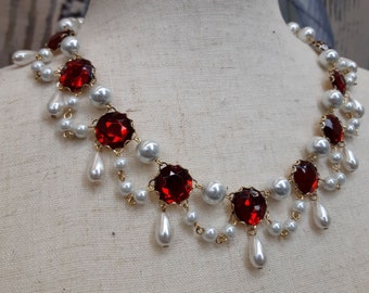 Può includere: Una collana in stile vintage con grandi pietre preziose rosse sfaccettate incastonate in metallo dorato. La collana è adornata con finte perle bianche e pendenti a goccia di perle, creando un design elegante e ornato.