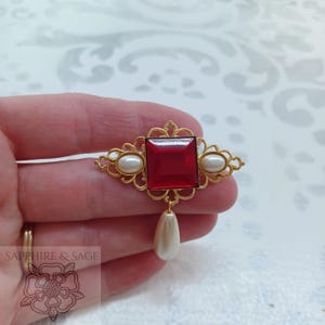 Puede incluir: Broche de estilo vintage con un marco dorado calado. Presenta una gran gema roja rubí cuadrada en el centro, flanqueada por dos perlas ovaladas. Una perla en forma de lágrima cuelga debajo. Un accesorio elegante.