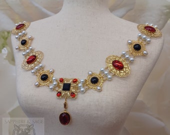 May include: A gold necklace with red and black gemstones, white pearls, and a filigree design. The necklace has a pendant with a red gemstone.